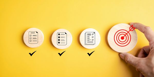 Wooden tokens with checklist icons and a target symbol being held by a hand on yellow background.