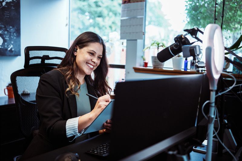 A Latina businesswoman in an executive position at a credit union sits at the desk in her office and uses a ring light while self-recording a financial literacy course for clients.