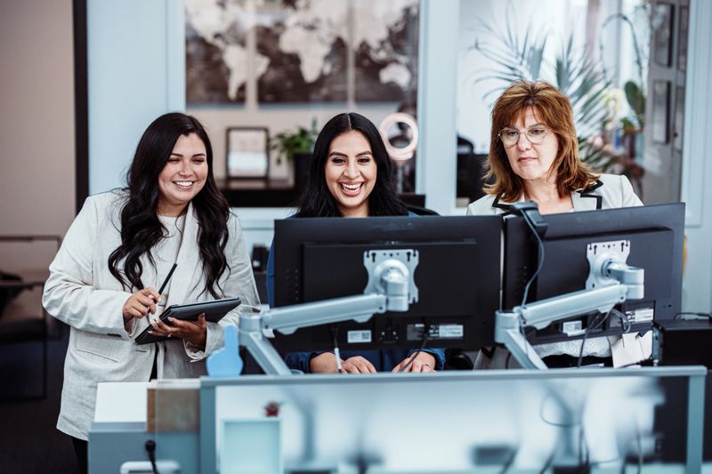Diverse group of female businesswomen of various ages work together at a computer station, collaborating on tasks in the modern credit union where they are employed.