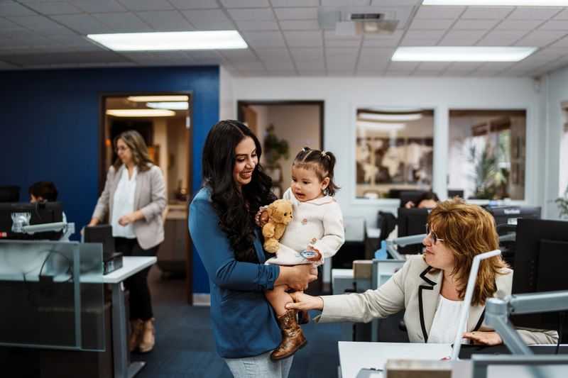 A Latin American woman holds her toddler daughter while chatting with a coworker at the credit union where she works, which supports family-friendly policies and flexible work arrangements.