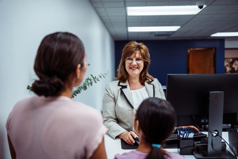 A female credit union representative smiles as she greets an unrecognizable female customer visiting the branch with her elementary-aged daughter.