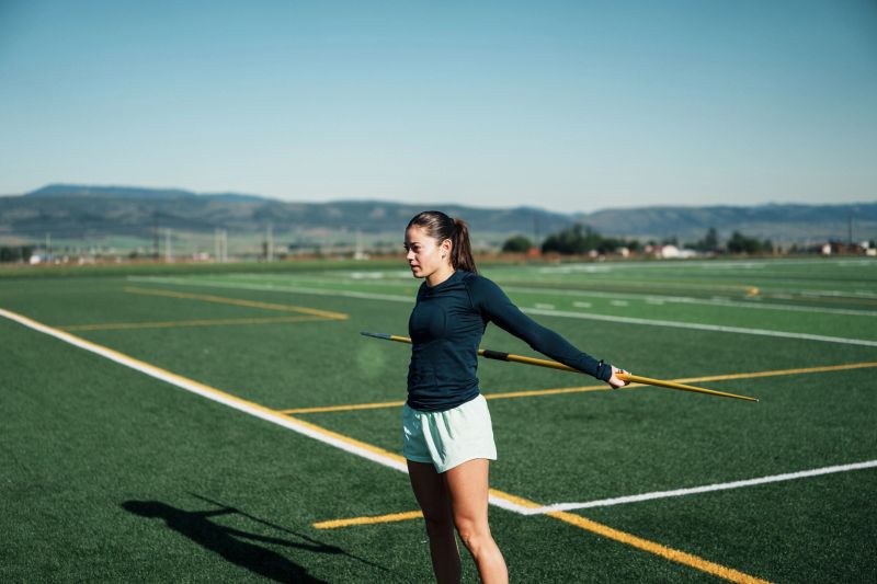 A female collegiate track and field athlete holds a javelin and performs arm stretches before training on the sports field at her former high school while home on summer break.