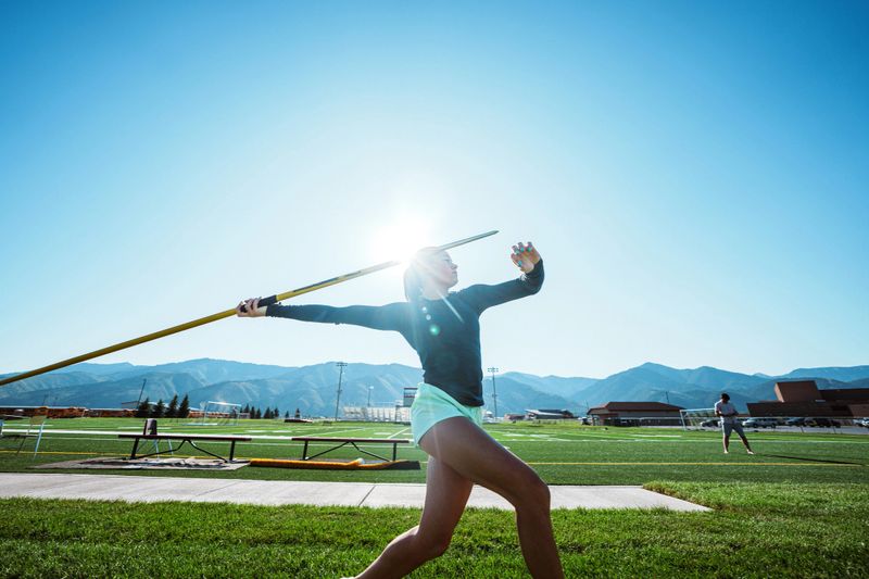 Profile view of a Latina collegiate heptathlete executing a powerful javelin throw on the field while her coach observes nearby during practice.
