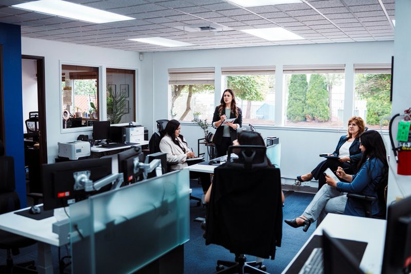 A Latin American woman in an executive role stands and leads a meeting with her team inside a modern credit union office.