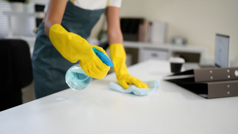 Young asian woman wearing yellow gloves and an apron, smiling while cleaning a white table with disinfectant spray and a blue cloth, promoting hygiene and cleanliness in her workspace.