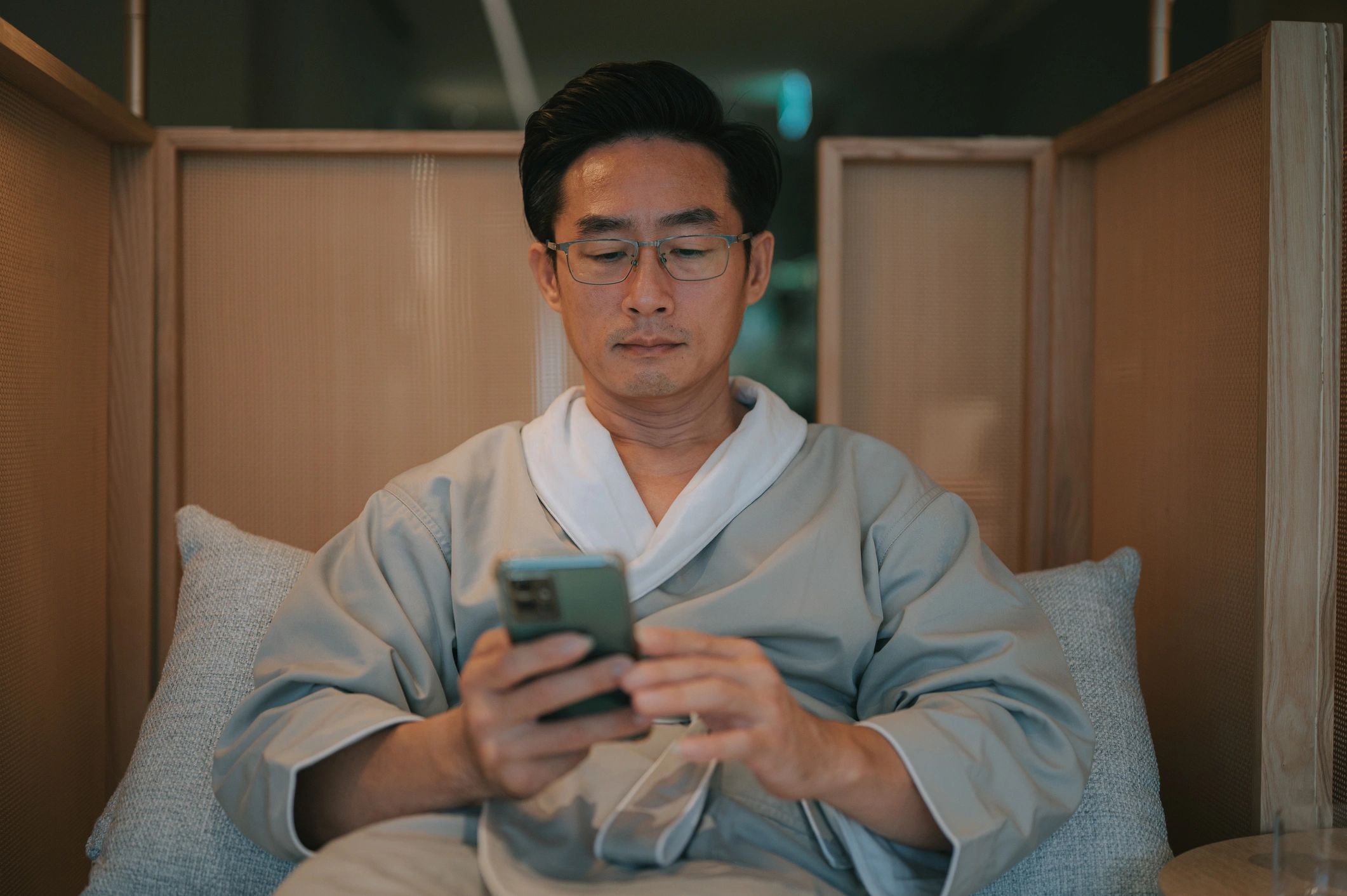 Front view of an Asian man wearing a spa robe sitting in a lounge chair and using a smartphone, symbolizing relaxation, digital lifestyle, and balance between technology and wellness. Kuala Lumpur.