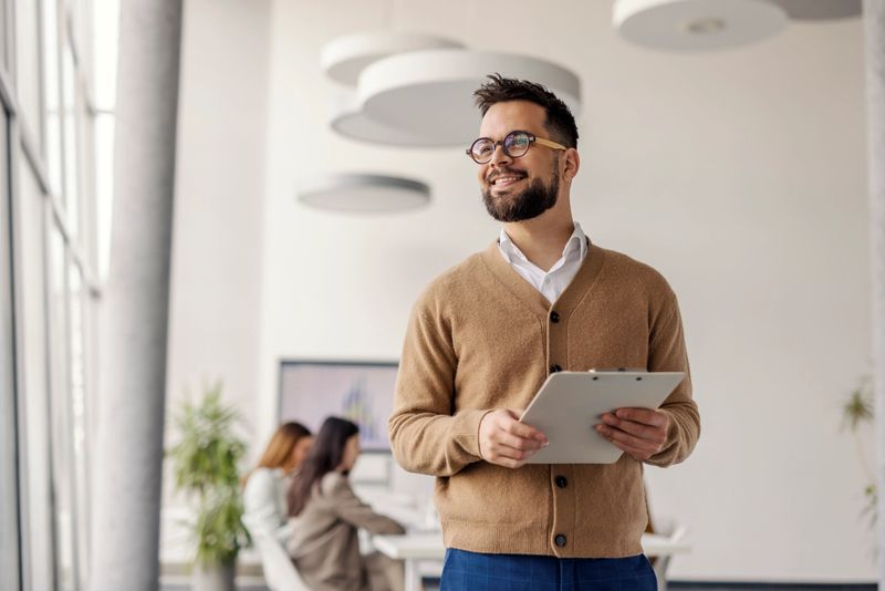 A young male professional with glasses and a beard stands confidently in a bright office holding a clipboard, with colleagues collaborating in the background.