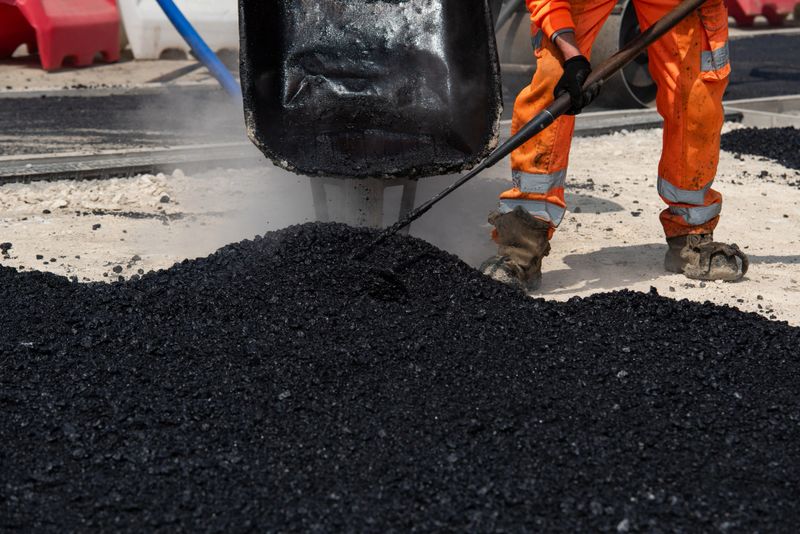 Team of road workers in bright orange uniforms repair road surface with wheelbarrow filled with hot asphalt