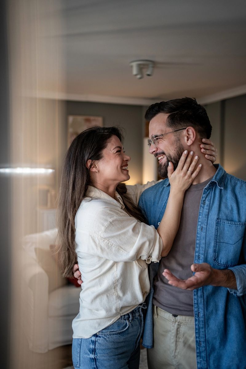 Mid adult couple is standing in their living room, embracing and smiling at each other, enjoying a quiet moment of love and connection in their cozy home