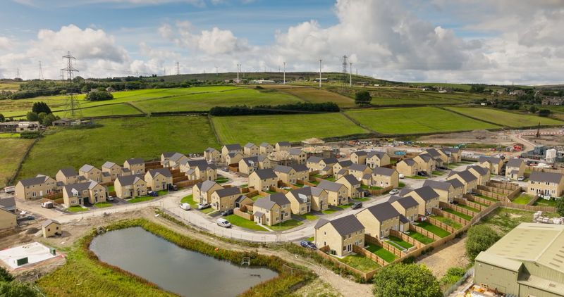 Drone shot of a modern residential housing estate under construction in Bradford, West Yorkshire. The development features rows of newly built homes with small gardens, surrounded by green agricultural fields and rolling countryside.  A small pond sits adjacent to the housing development, with construction equipment and materials visible throughout the site.
