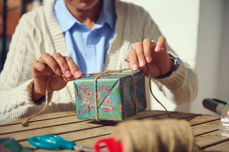 Close-up of hands tying string around a beautifully wrapped gift, surrounded by crafting tools, in a warm and sunny environment. Suggests creativity, care, and thoughtfulness in gift preparation.