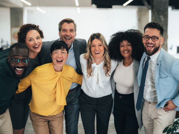 Seven mixed aged & race professionals putting their arms on each other's shoulders smiling.