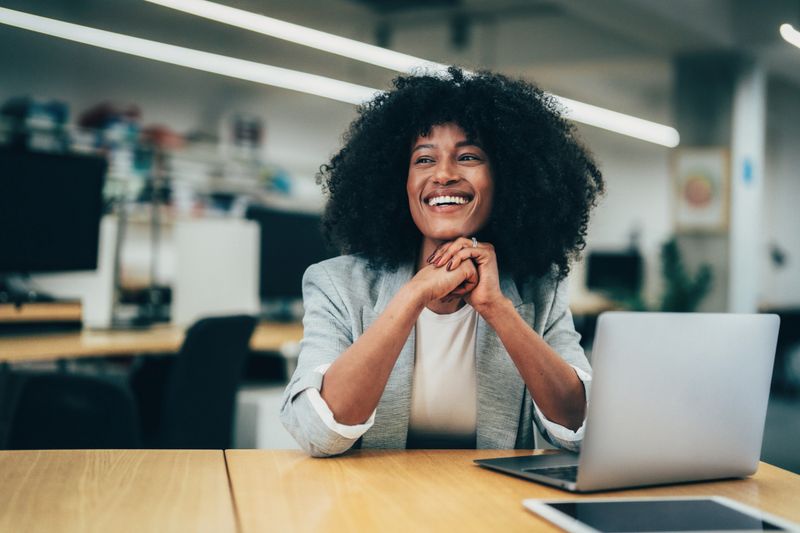 Confident and friendly professional woman sitting at a desk in a modern office. A laptop is open on the desk in front of her, indicating she may be engaged in work, a video call, or some form of digital task.