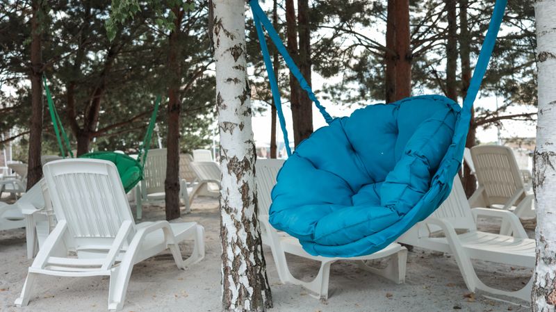 A vibrant blue hanging chair is the focal point, surrounded by white lounge chairs and trees, creating a tranquil beach setting. The scene evokes relaxation and leisure.