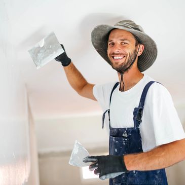 Smiling worker plastering a wall indoors wearing a hat and gloves.