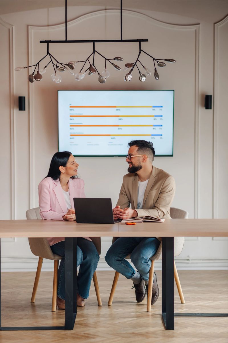 Two business colleagues are sitting at a table, discussing work and looking at a laptop, with a graph displayed on a screen behind them