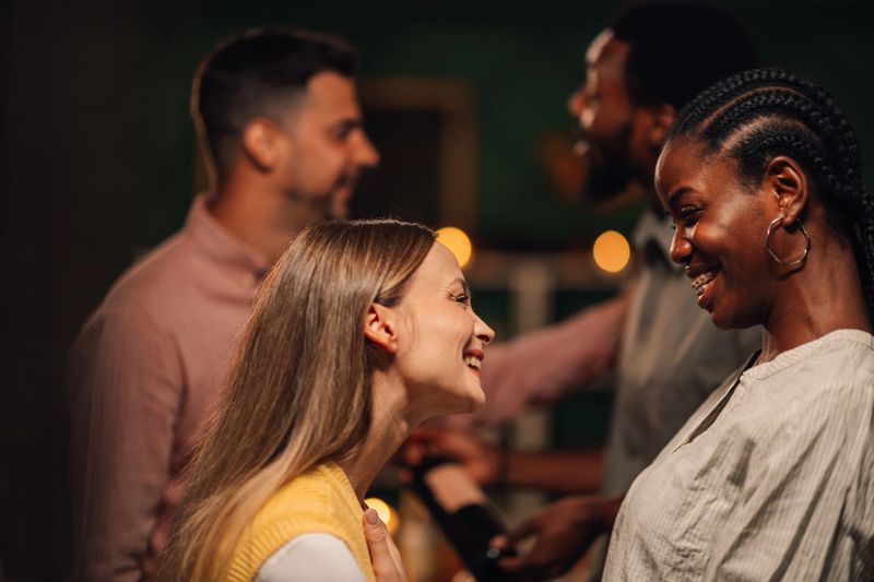Two women laughing and chatting joyfully at a lively party, surrounded by friends enjoying the festive atmosphere in the background