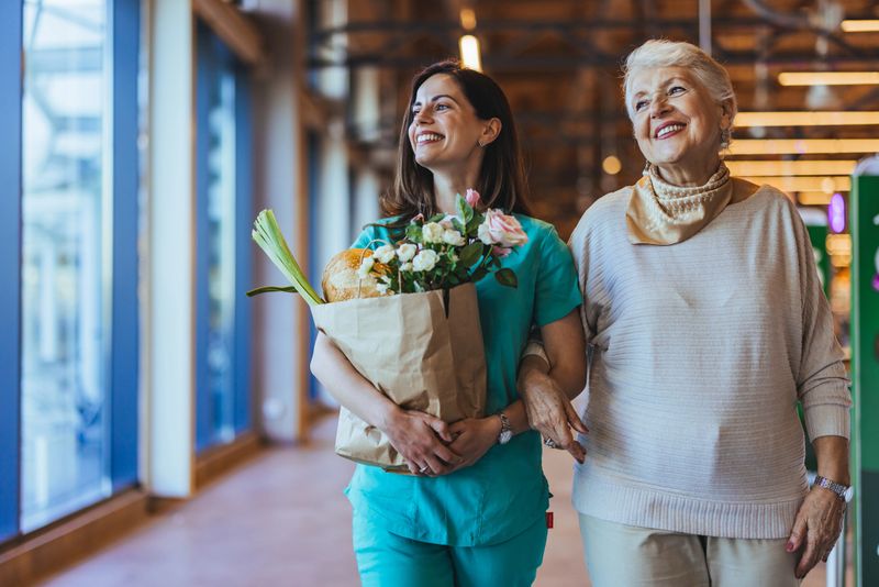 A healthcare worker assists an elderly woman in a market, holding a paper bag of groceries and flowers. They smile as they walk together, sharing a joyful moment.