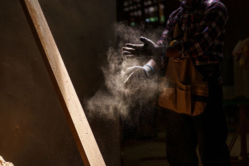 Carpenter brushes sawdust off his hands after finishing a woodworking task. Sunlight highlights the airborne dust, creating a beautiful moment that reflects craftsmanship, and dedication of labor.