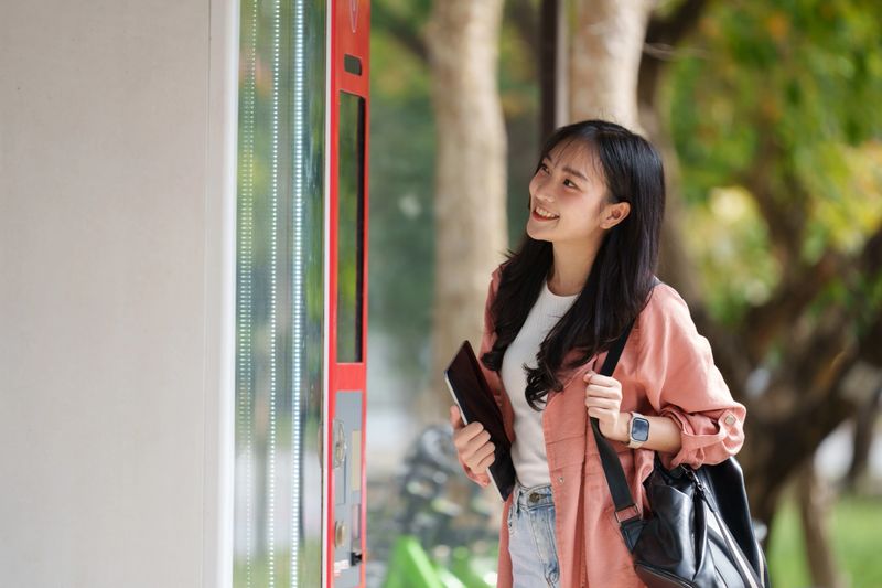 Smiling student is using a modern vending machine outdoors, holding a tablet and wearing a smartwatch and backpack, enjoying a convenient and cashless purchase experience