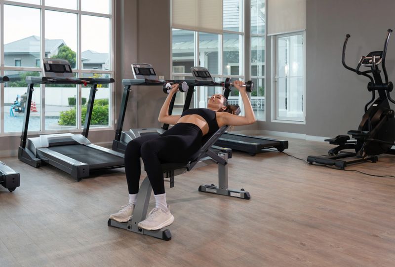 Young woman in sport clothing lean on the bench in a contemporary gym, perform a incline dumbbell press. Natural light stream through large window, illuminating the fitness space.