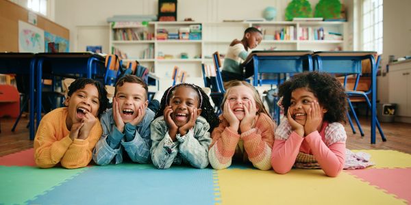 Five happy children lying on a colorful mat in a classroom.