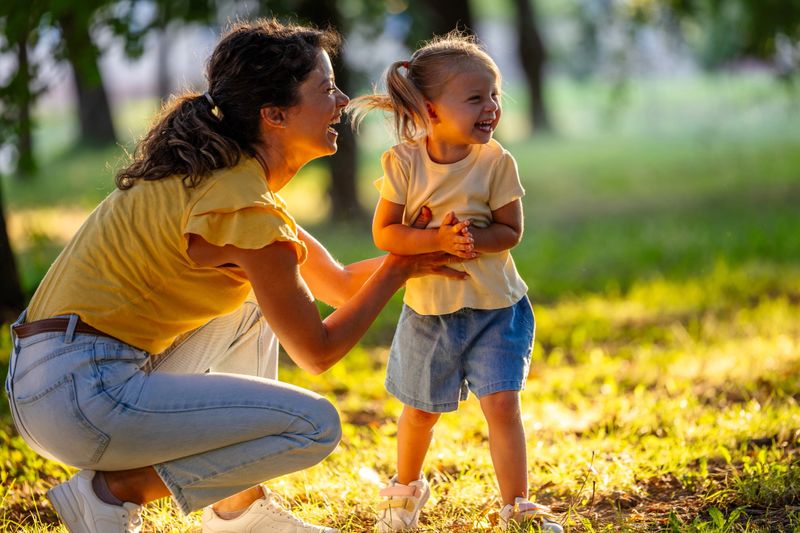 Woman and her young daughter enjoy laughter together in sunlit park surrounded by trees during late afternoon
