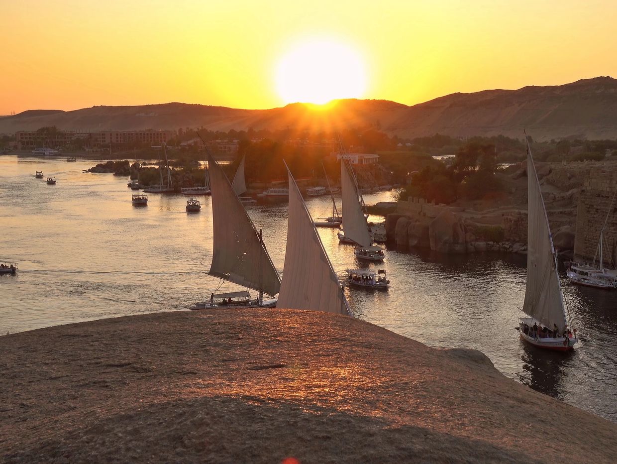Sailboats glide on a river at sunset, with hills in the background.