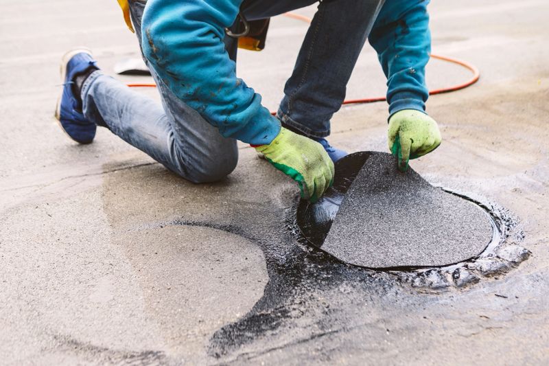 Roofer repairing a damaged section of a roof. Worker kneels on a wet, flat rooftop to apply a circular patch. Their gloved hands press the black material into place, sealing a roof