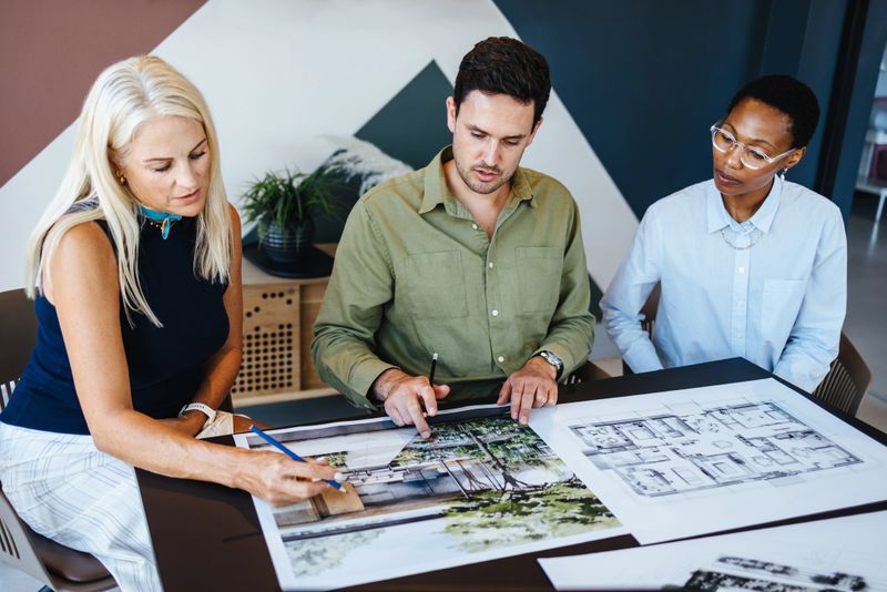 A team of professionals of different genders and age groups engaging in a design discussion, examining architectural plans in an office space, fostering collaboration and innovative brainstorming.
