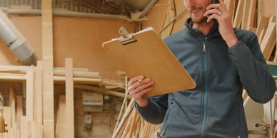 A man talks on phone while holding a clipboard in a woodworking shop.