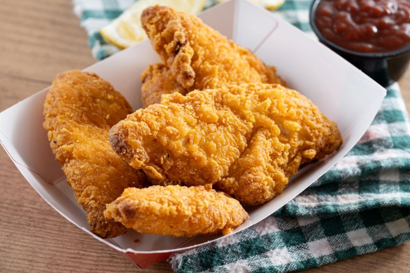 Close-up of golden crispy fried chicken tenders served in a red and white paper tray on wooden table.
