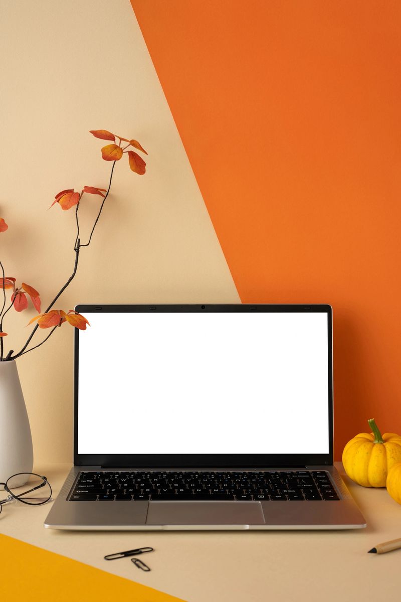 Stylish vertical workspace featuring a laptop, autumn leaves, and decor on a vibrant orange desk