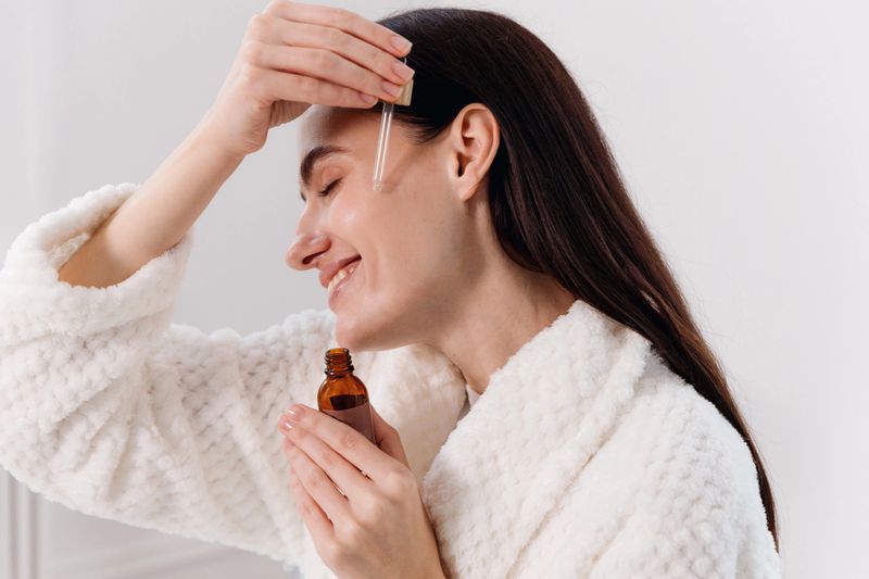In modern bathroom, young woman in a bathrobe using glass dropper, applies serum oil with vitamin c to her face as part of her anti aging morning skincare routine, embracing self-care and wellness.