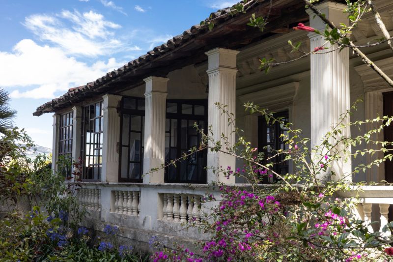 Elegant colonial hacienda in Ecuador featuring white columns, wooden balcony windows, tiled roof, and a colorful flowering garden that highlights its historic charm.