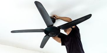 A person installing a black ceiling fan with five blades.