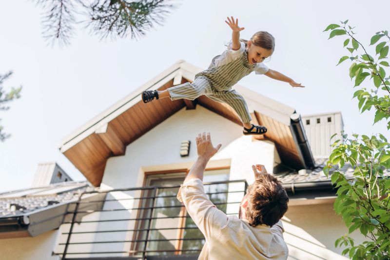 Father playfully throwing his daughter in the air, enjoying a sunny day in front of their modern home