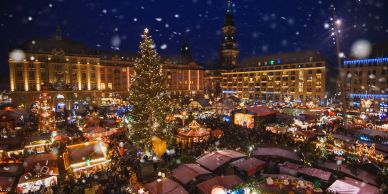 Festive Christmas market at night with a large Christmas tree and falling snow.