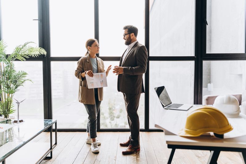 Two architects engaging in a detailed discussion by the window of a modern office, collaborating on innovative project plans