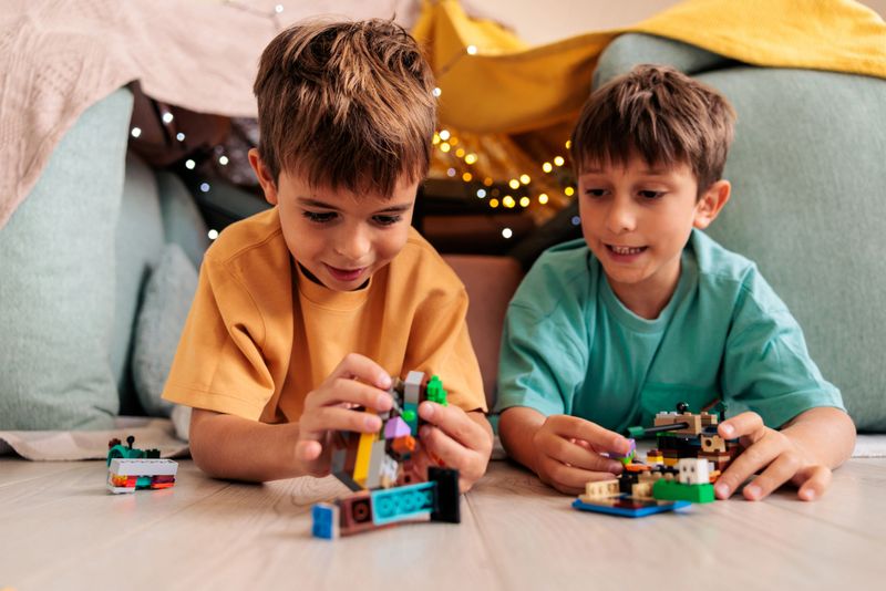 Two young boys lie on their stomachs inside a cozy blanket fort, playing with toys and enjoying their indoor space. The scene captures fun, creativity, and a warm, playful atmosphere.