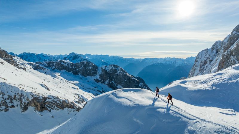 Two adventurers are skillfully navigating a snow-covered alpine landscape at sunrise, featuring breathtaking views of rugged mountain peaks and a clear sky. They are equipped with skis and winter gear, enjoying the serene and challenging environment.