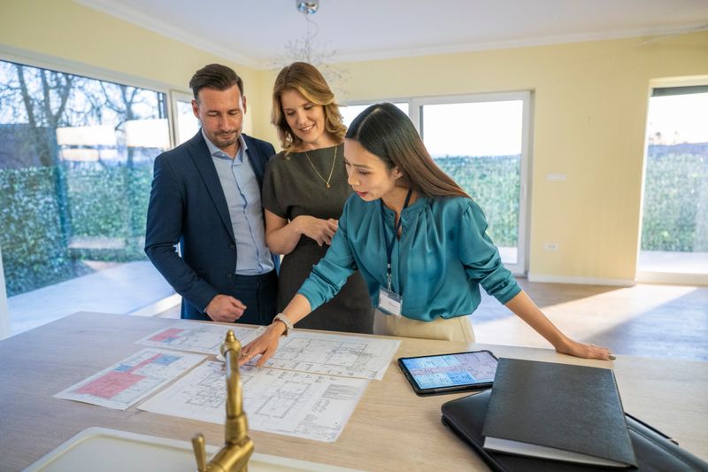 A mid adult Asian female real estate agent is discussing house blueprints with a Caucasian couple in a bright room. She is pointing at the plans on a table featuring natural light from large windows.