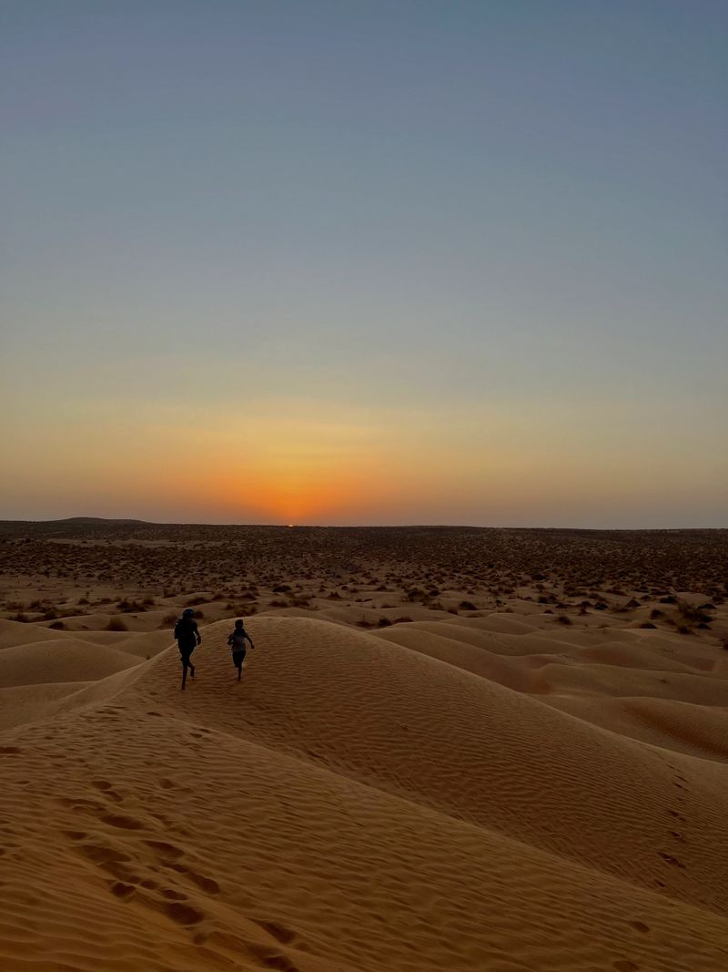 Two children playfully run across the golden sand dunes of Douz, Tunisia, enjoying the tranquil evening as the sun sets in the background, creating a serene atmosphere.