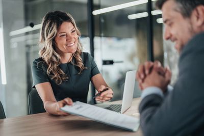 A woman smiling while handing a clipboard to a man in a business setting.