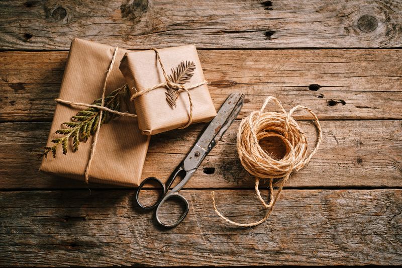 Two beautifully wrapped gifts with brown paper and twine are placed on a rustic wooden table. Scissors and natural decorations add charm to the scene.