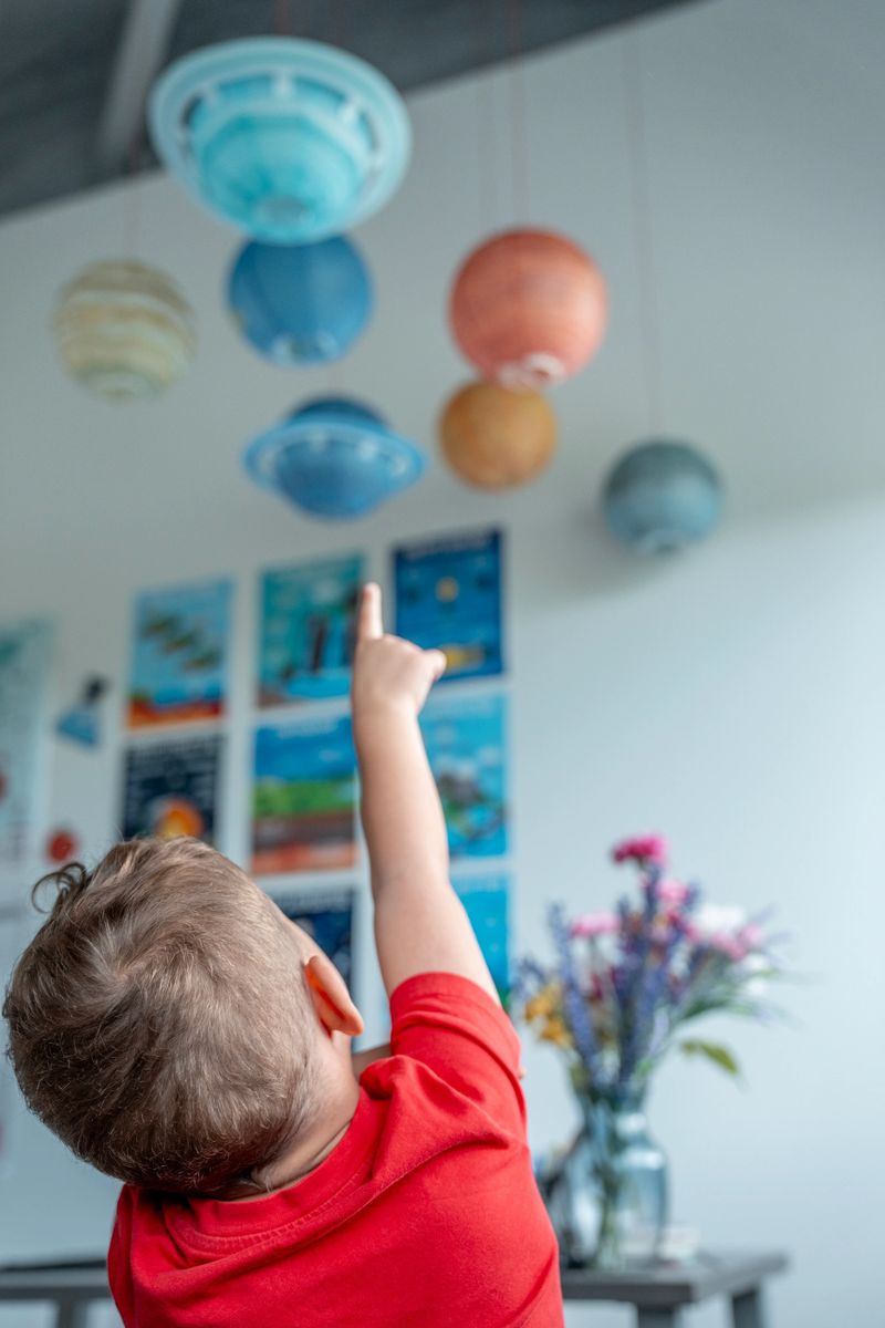 A child dressed in a red shirt points enthusiastically at vibrant planet models hanging from above, showcasing curiosity and exploration in a colorful and educational setting.