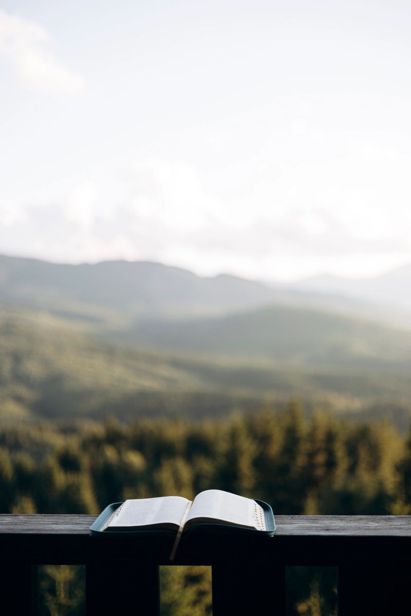 An open Bible rests on a wooden balcony railing overlooking a peaceful mountain landscape. A serene scene of faith, reflection, and connection with nature.