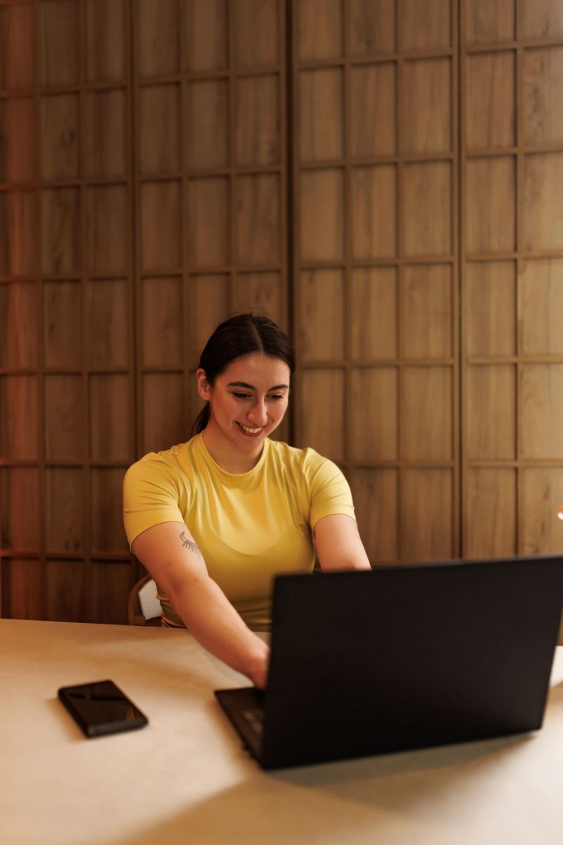 A woman in a yellow shirt is focused on her laptop while seated at a wooden table. She has a cheerful expression, and a smartphone is placed next to her.