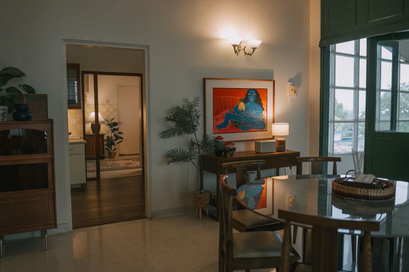 Interior view of a dining room featuring a round glass table, wooden furniture, wall artwork, and warm lamp lighting, with doorway view into the kitchen