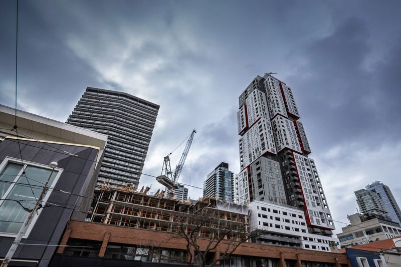 This urban scene documents the transformation of Queen Street in downtown Toronto. Cranes pivot above a busy construction site where new condominium towers are being formed in concrete and steel. Surrounding high-rises create a dense skyline, while streetcar wires and low-rise fronts anchor the street level. The picture communicates real estate development, investment and the pressures of housing demand in Canada's largest city. It can illustrate property markets, architecture, planning, mixed-use projects, and the continuous cycle of redevelopment that reshapes neighborhoods across the GTA.